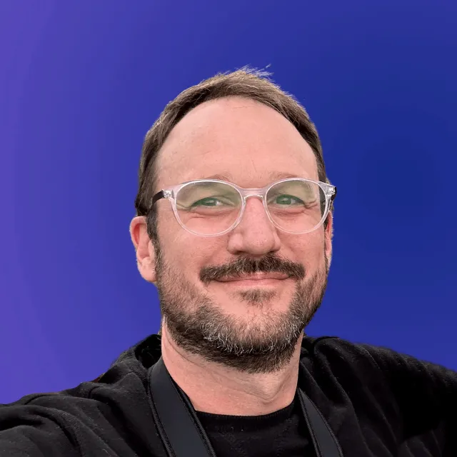 Jakes van Eeden, smiling, in clear-framed glasses and a black top, against a blue background.
